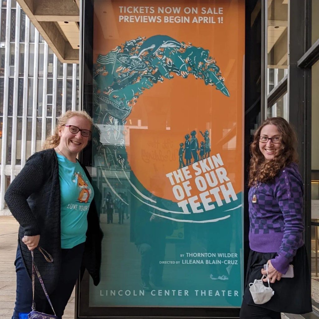Two women, smiling, standing on either side of a poster for The Skin of Our Teeth